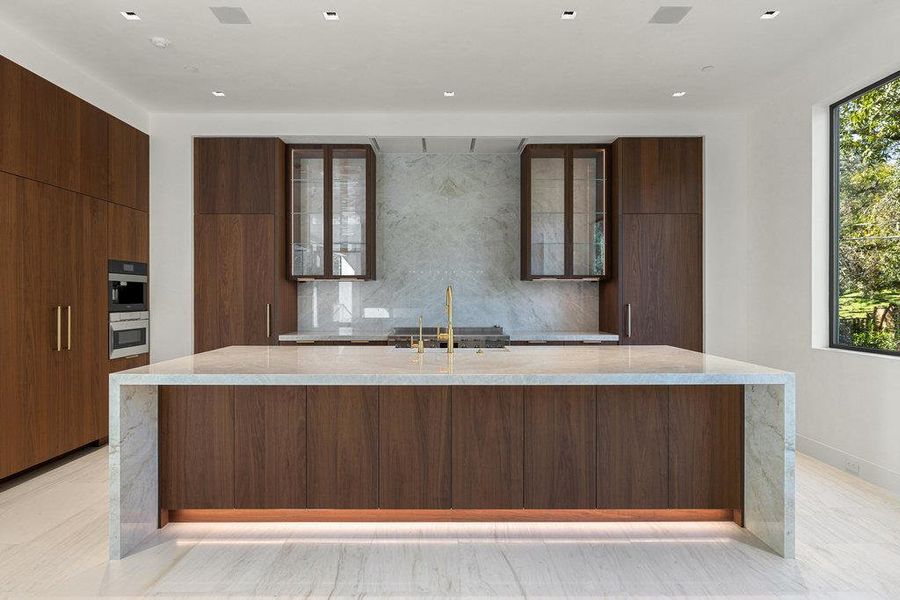 Kitchen featuring backsplash, light stone countertops, modern cabinets, a spacious island, and glass insert cabinets