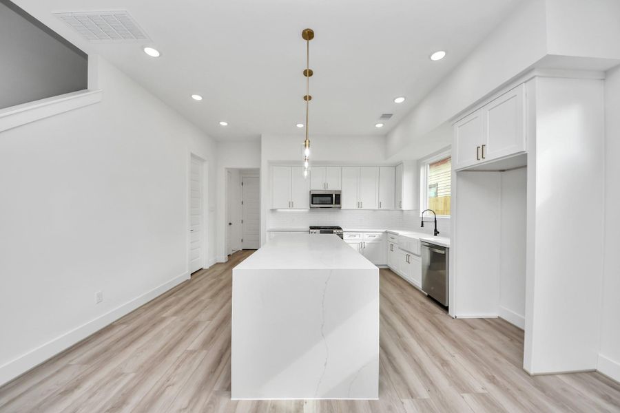 This modern kitchen features sleek white cabinetry, a large island with marble-like countertops, stainless steel appliances, and pendant lighting. The open layout with light wood flooring creates a bright and inviting space.
