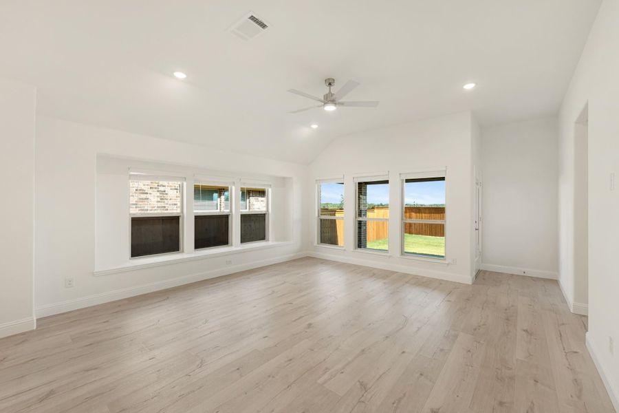 Empty living room with vaulted ceiling, ceiling fan, light wood floors, and multiple windows overlooking backyard