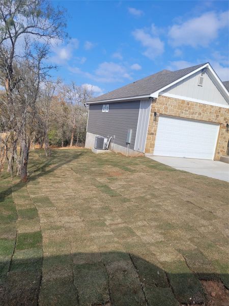 View of side of home featuring a lawn, driveway, stone siding, a shingled roof, and an attached garage