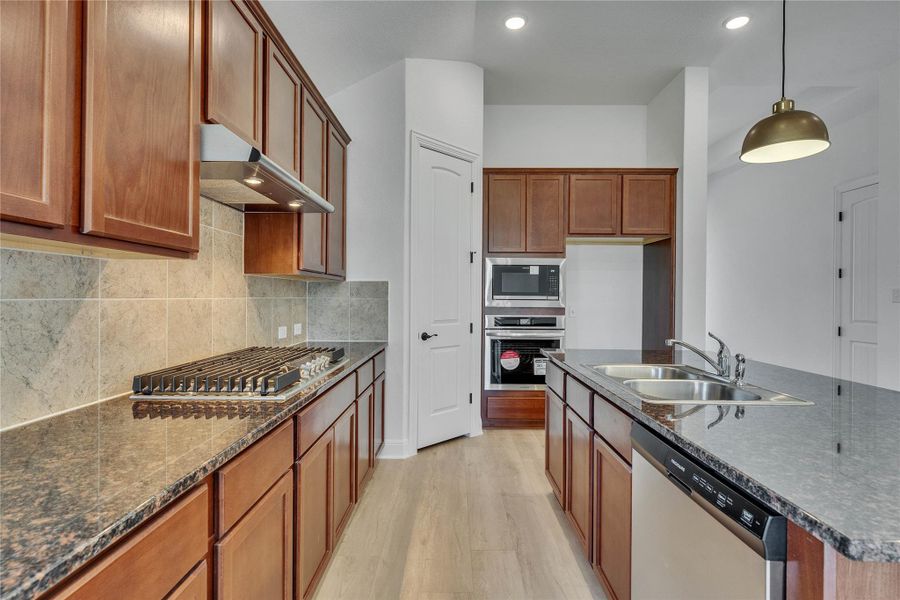 Kitchen featuring brown cabinetry, stainless steel appliances, light wood finished floors, a center island with sink, and under cabinet range hood Kitchen featuring brown cabinetry, stainless steel appliances, light wood finished floors, a center island with sink, and under cabinet range hood