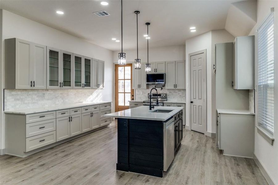 Kitchen featuring hanging light fixtures, light wood finished floors, a kitchen island with sink, backsplash, and gray cabinets Kitchen featuring hanging light fixtures, light wood finished floors, a kitchen island with sink, backsplash, and gray cabinets