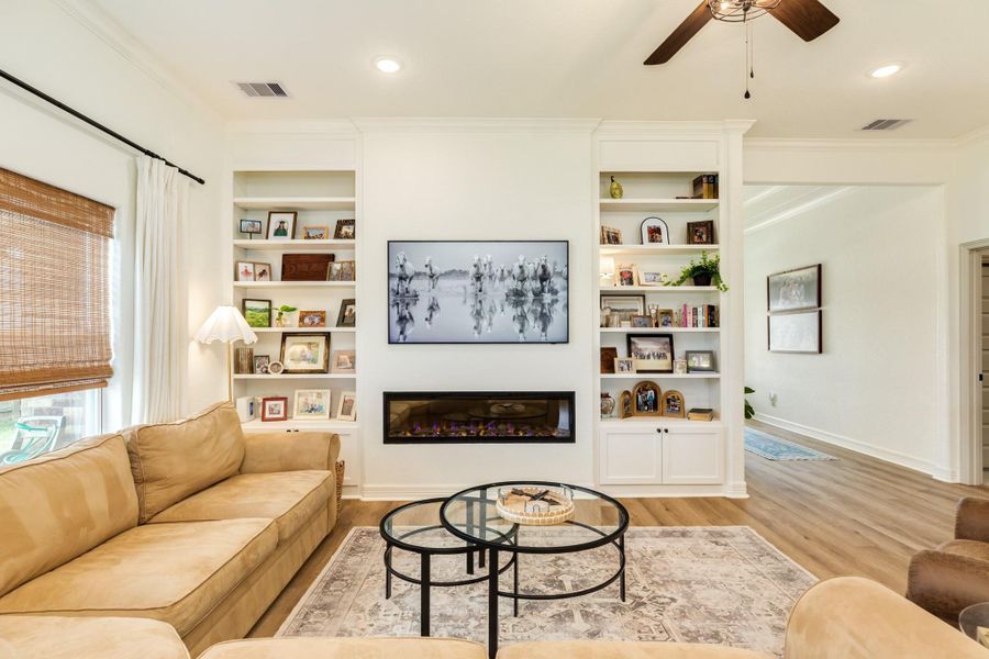 This inviting living room features, custom built-in shelves flanking a sleek fireplace. The space is accented by warm wood look flooring, and ample natural light from large windows with bamboo styled blinds.