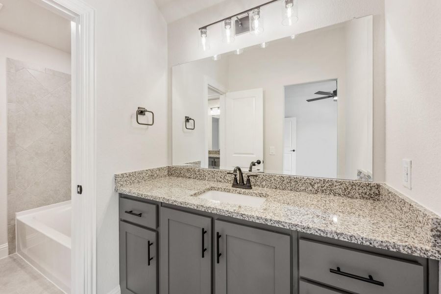 Bathroom with gray vanity, granite countertop, large mirror, and tiled tub-shower combo visible in background.
