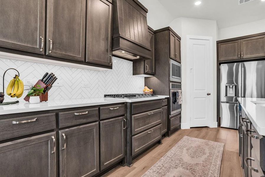 Kitchen featuring a substantial island with breakfast bar, undermount sink with gooseneck brushed nickel faucet, wood-finish flooring, dark wood cabinetry, a white herringbone tile backsplash, walk-in pantry and stainless steel appliances including a propane gas cooktop, electric built-in oven and microwave open to living and breakfast rooms