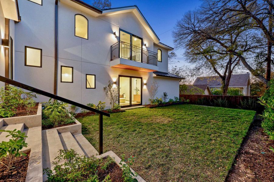 White stucco exterior featuring a second-story balcony with iron railing, modern wall-mounted lighting, and a manicured lawn