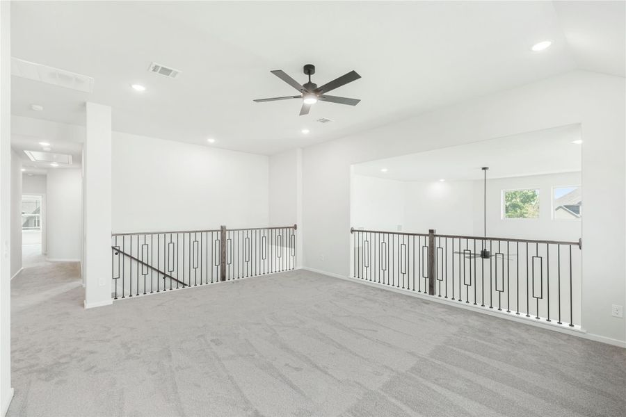 Open upstairs loft with gray carpet, ceiling fan, and black metal stair railings on two sides Open upstairs loft with gray carpet, ceiling fan, and black metal stair railings on two sides