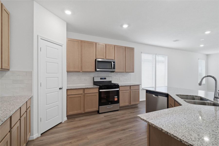 This spacious kitchen boasts soaring ceilings, Woodmont Dakota cabinets in a Barley stain, Granite Iberian White countertops, stainless steel Whirlpool appliances, a modern Daltile backsplash, recessed lighting, an extended breakfast bar countertop, and a pantry—all overlooking the expansive family room.