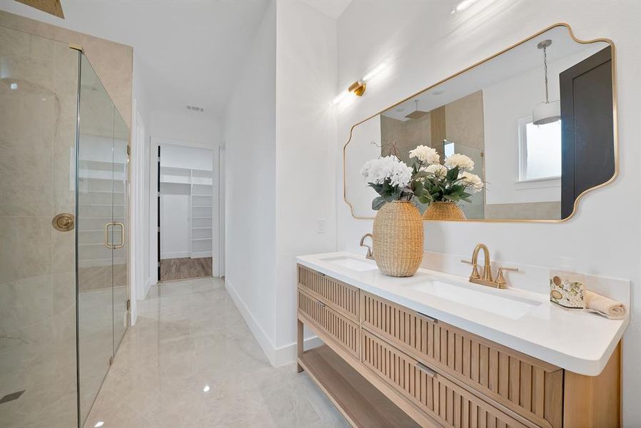 The bathroom features a double vanity with a light wood finish and brass fixtures, a frameless shower with a brass handle, and light-colored tile flooring