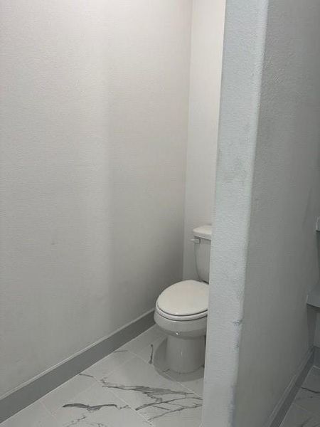 This is a compact, modern bathroom featuring a white toilet and sleek, marble-patterned tile flooring, captured from an angle that shows the clean lines and neutral color palette.