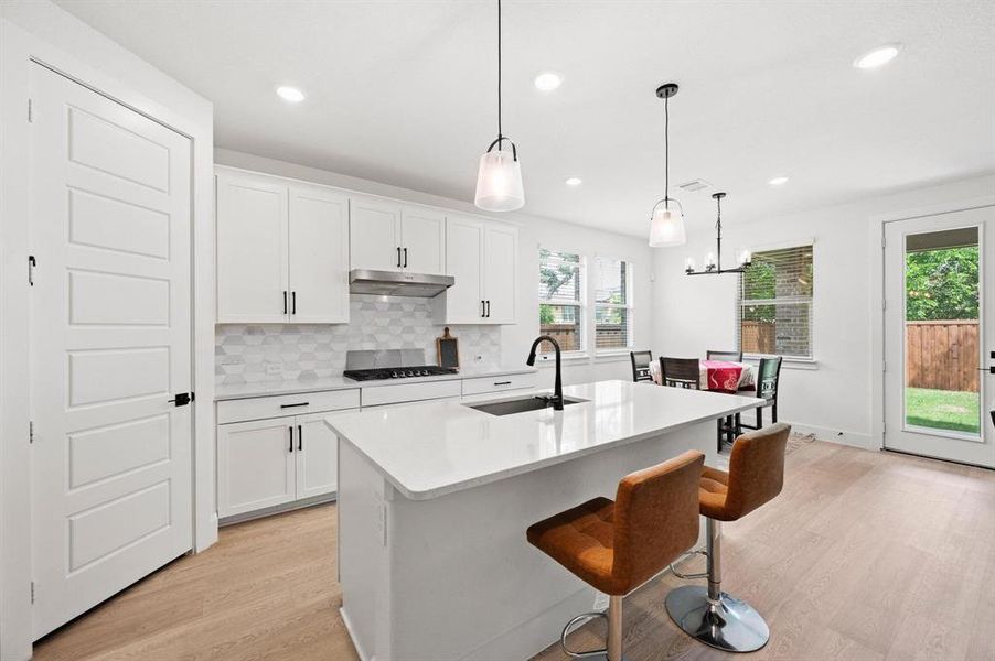 Modern kitchen featuring an expansive island with a pristine white countertop, light wood-finish flooring, a white hex-tile backsplash, sleek stainless steel appliances, and abundant natural light Modern kitchen featuring an expansive island with a pristine white countertop, light wood-finish flooring, a white hex-tile backsplash, sleek stainless steel appliances, and abundant natural light