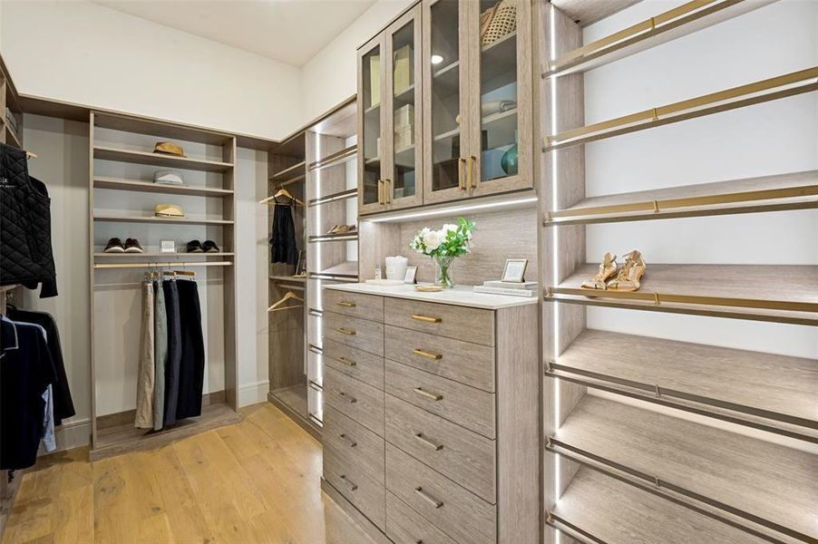 Custom closet system featuring wood-finish cabinetry, integrated LED lighting, glass-front upper cabinets, a central island with drawer storage, and warm wood flooring