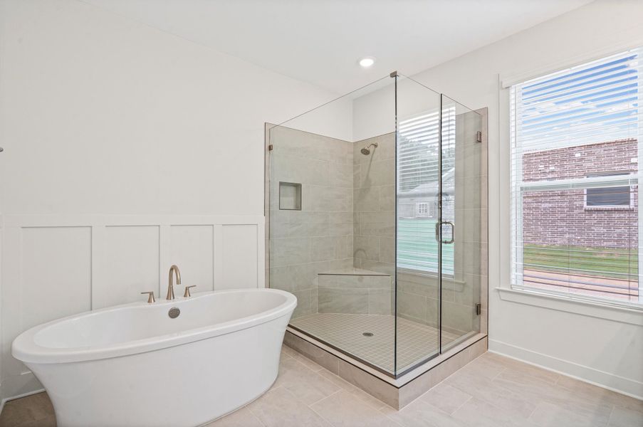 Freestanding soaking tub with brushed brass fixtures, glass-enclosed shower with a tiled bench, and large window with blinds Freestanding soaking tub with brushed brass fixtures, glass-enclosed shower with a tiled bench, and large window with blinds