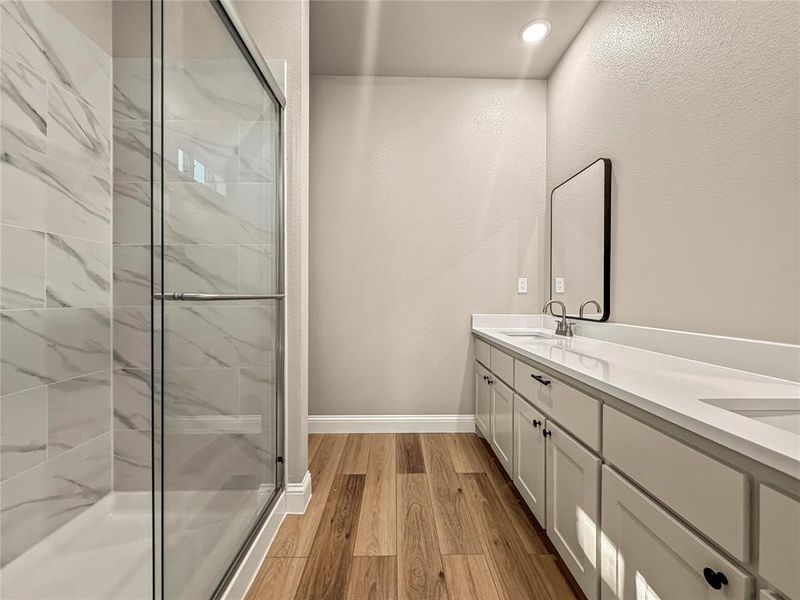 Spacious bathroom featuring a frameless glass shower with white marble-look tile, wood-finish flooring, a double vanity with white countertops, and white shaker-style cabinetry with matte black hardware Spacious bathroom featuring a frameless glass shower with white marble-look tile, wood-finish flooring, a double vanity with white countertops, and white shaker-style cabinetry with matte black hardware