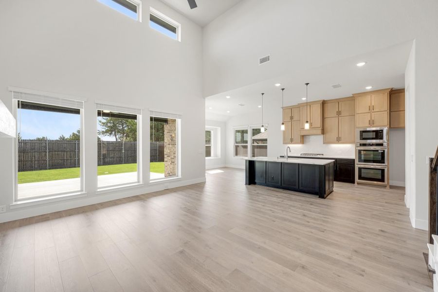 Open-concept living area with high ceilings, light wood floors, and kitchen with dark island and tan cabinets Open-concept living area with high ceilings, light wood floors, and kitchen with dark island and tan cabinets