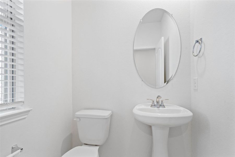 The powder room on the first floor includes a pedestal sink, mirror, toilet, and window with privacy blinds. The powder room on the first floor includes a pedestal sink, mirror, toilet, and window with privacy blinds.