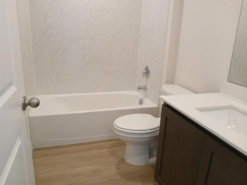Full bathroom featuring a white tub with integrated shower, white toilet, wood-finish flooring, a dark wood-finish vanity, and a white countertop with an integrated sink Full bathroom featuring a white tub with integrated shower, white toilet, wood-finish flooring, a dark wood-finish vanity, and a white countertop with an integrated sink