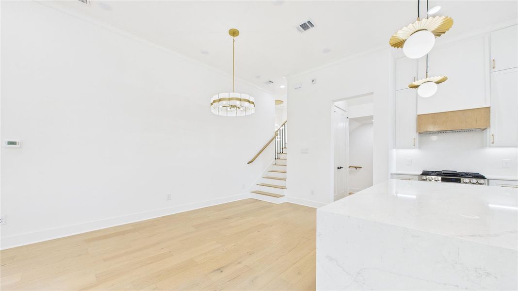 Bright and modern open-concept space featuring light wood floors, white walls, and elegant gold-accented light fixtures. The kitchen includes a sleek marble island and high-end appliances. A staircase leads to the upper level, adding a touch of sophistication.