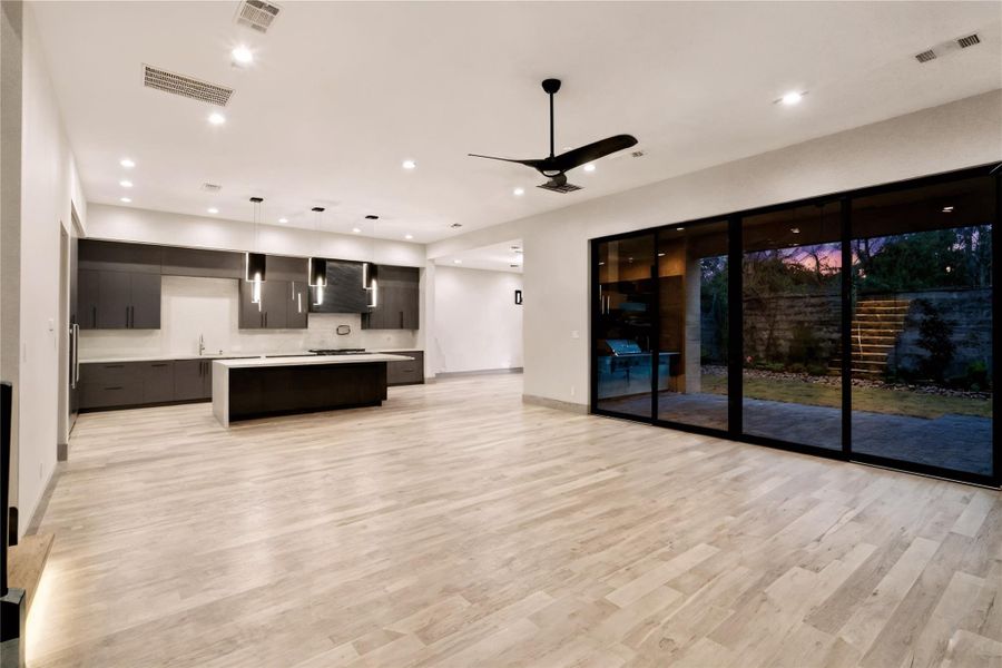 This modern, open-concept living space features a sleek kitchen with dark cabinetry, a large island, and pendant lighting. The room is bright and airy with light wood flooring, recessed lighting, and expansive sliding glass doors that open to a landscaped outdoor area, ideal for seamless indoor-outdoor living.