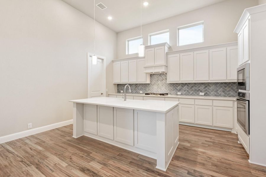 Kitchen with white cabinets, large island, gray herringbone backsplash, and wood-look floors with high ceilings