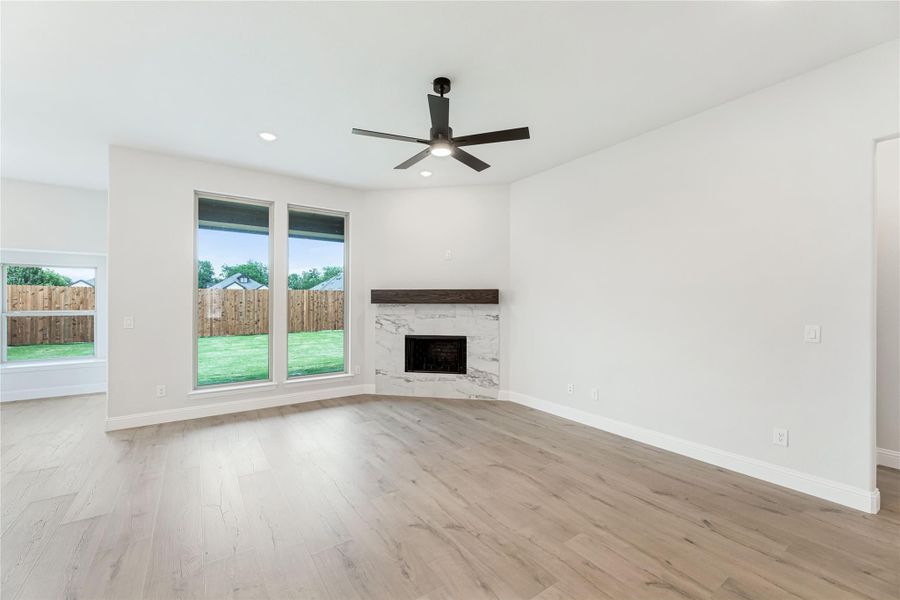 Open living room with light wood floors, stone fireplace, ceiling fan, and large backyard windows