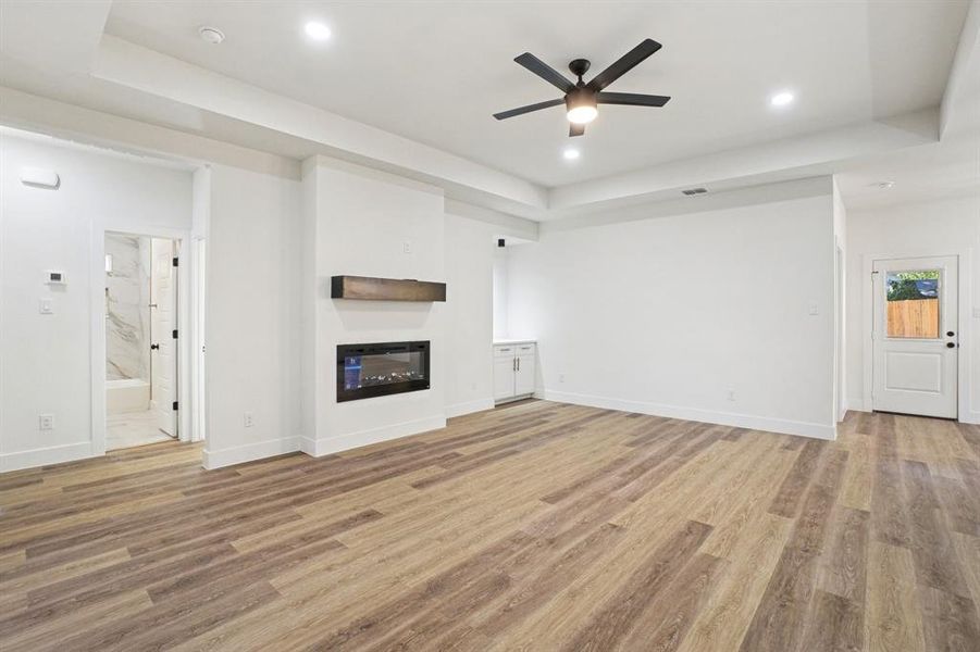 Unfurnished living room featuring a tray ceiling, a glass covered fireplace, ceiling fan, light wood-style floors, and recessed lighting Unfurnished living room featuring a tray ceiling, a glass covered fireplace, ceiling fan, light wood-style floors, and recessed lighting