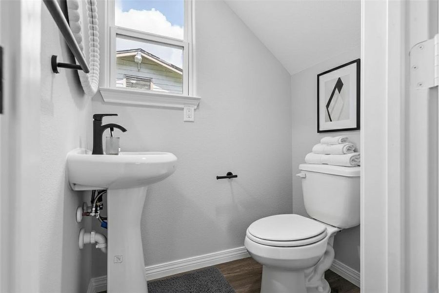 This is a compact, modern half-bathroom featuring a pedestal sink, a toilet, and a small window for natural light. **Photo has been staged.**