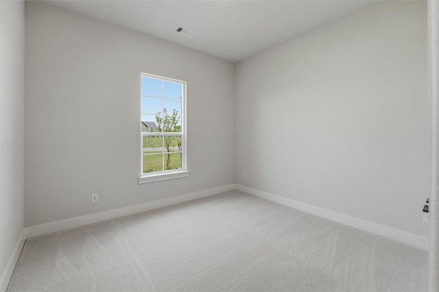 Room with light gray walls and plush gray carpet, featuring a window with a view of a grassy outdoor area and a distant roofline