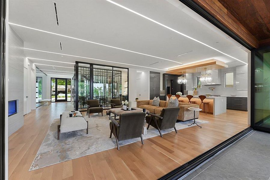 Open-concept living space featuring wood-finish flooring, linear ceiling lighting, a built-in fireplace, a glass-enclosed feature wall, and a kitchen with an island and pendant lighting