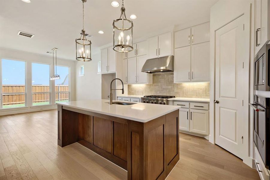 Kitchen with light wood-type flooring, tasteful backsplash, a kitchen island with sink, stainless steel appliances, and a chandelier