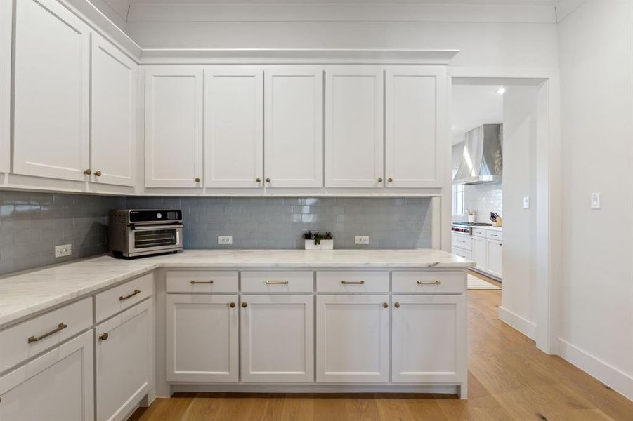 Kitchen with light wood-type flooring, wall chimney range hood, white cabinets, and backsplash Kitchen with light wood-type flooring, wall chimney range hood, white cabinets, and backsplash
