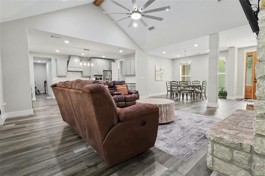 Spacious open-concept interior featuring vaulted ceilings with a beam accent, wood-finish flooring, a stone fireplace, and recessed lighting