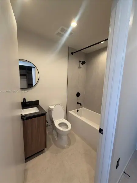 Bathroom featuring a wood-finish vanity with a dark stone countertop, circular mirror, and matte black hardware including a shower-tub faucet assembly with full-height tile surround