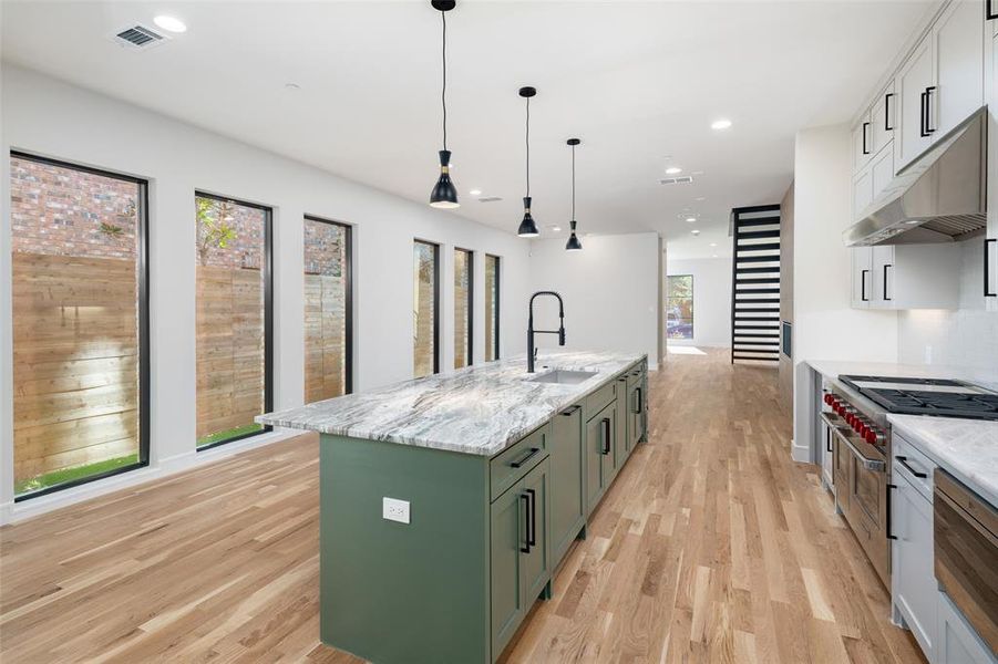 Kitchen with light stone counters, sink, a center island with sink, green cabinetry, and hanging light fixtures