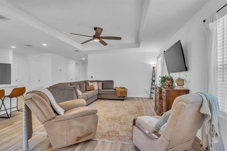 Living room with a raised ceiling, light wood-style flooring, ceiling fan, recessed lighting, and crown molding