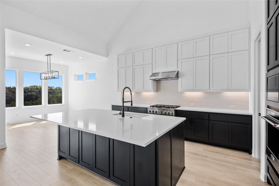 Dual tone kitchen with two tone cabinetry, light wood-style flooring, hanging light fixtures, a kitchen island with sink, and decorative backsplash