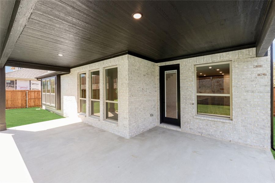 Covered back patio with concrete floor, dark ceiling, and light brick exterior wall with large windows and door. Covered back patio with concrete floor, dark ceiling, and light brick exterior wall with large windows and door.