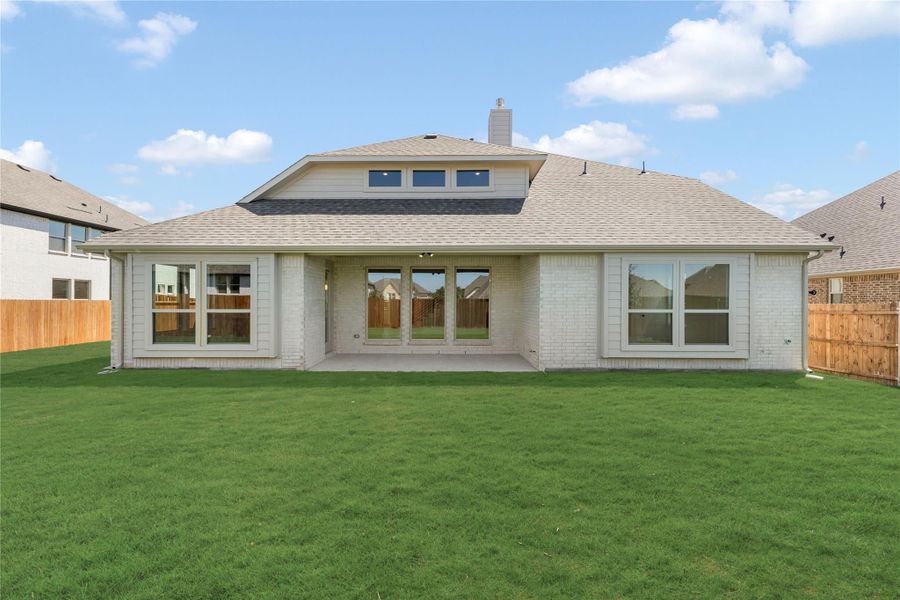 Rear view of single-story brick home with covered patio, large windows, and fenced backyard with green lawn.
