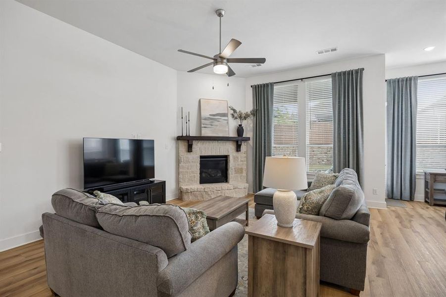 Living area featuring wood-finish flooring, a stone-clad fireplace with a dark wood mantel, multiple windows with blinds and drapery, a ceiling fan, and recessed lighting
