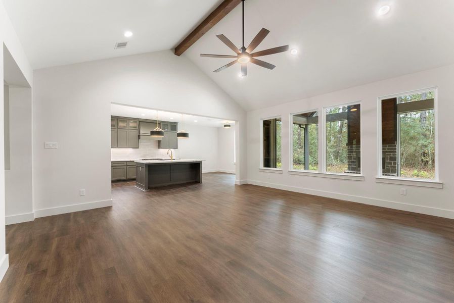 Another view of this spacious open-concept living area features high ceilings, large windows with forest views, and stylish wood flooring. The modern kitchen includes dark cabinetry, an island, and pendant lighting, creating an ideal setting for entertaining.