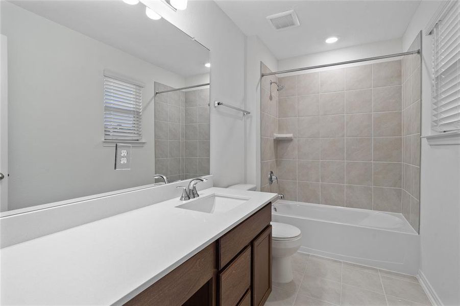 Secondary bath features tile flooring, bath/shower combo with tile surround, dark stained wood cabinets, beautiful light countertops, mirror, dark, sleek fixtures and modern finishes.