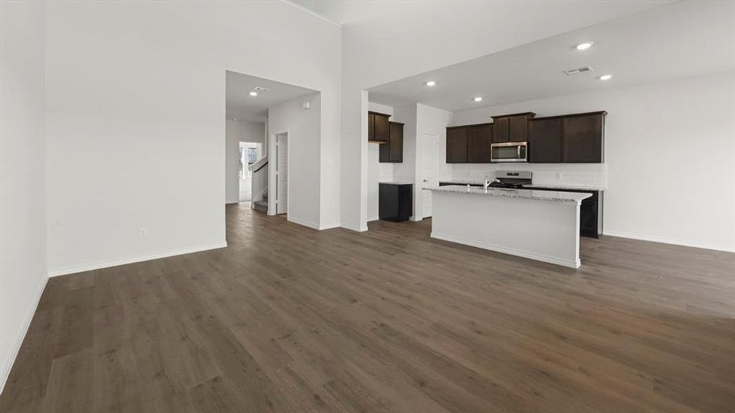 Expansive open-concept living area featuring wood-finish flooring, recessed lighting, a kitchen island with seating, dark wood cabinetry, and stainless steel appliances Expansive open-concept living area featuring wood-finish flooring, recessed lighting, a kitchen island with seating, dark wood cabinetry, and stainless steel appliances