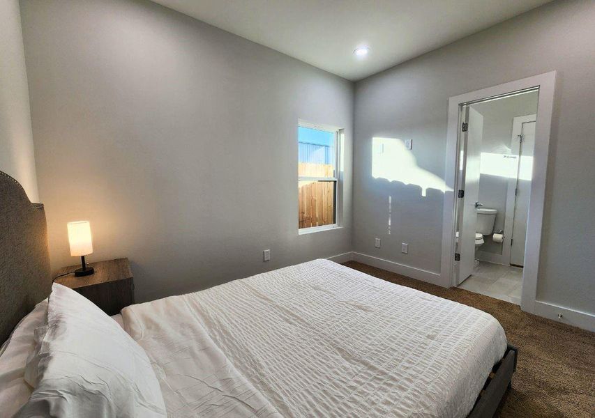 Cozy bedroom with neutral tones, featuring a comfortable bed, bedside table with a lamp, and a window letting in natural light. Adjoining bathroom with modern fixtures. Cozy bedroom with neutral tones, featuring a comfortable bed, bedside table with a lamp, and a window letting in natural light. Adjoining bathroom with modern fixtures.
