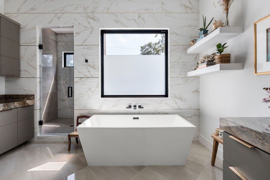 The freestanding soaking tub is set against a marble tile wall with a large picture window above, creating a bright and relaxing focal point within the primary bathroom. To the left is a huge walk in- shower with glass enclosure, 2 shower heads and corner bench