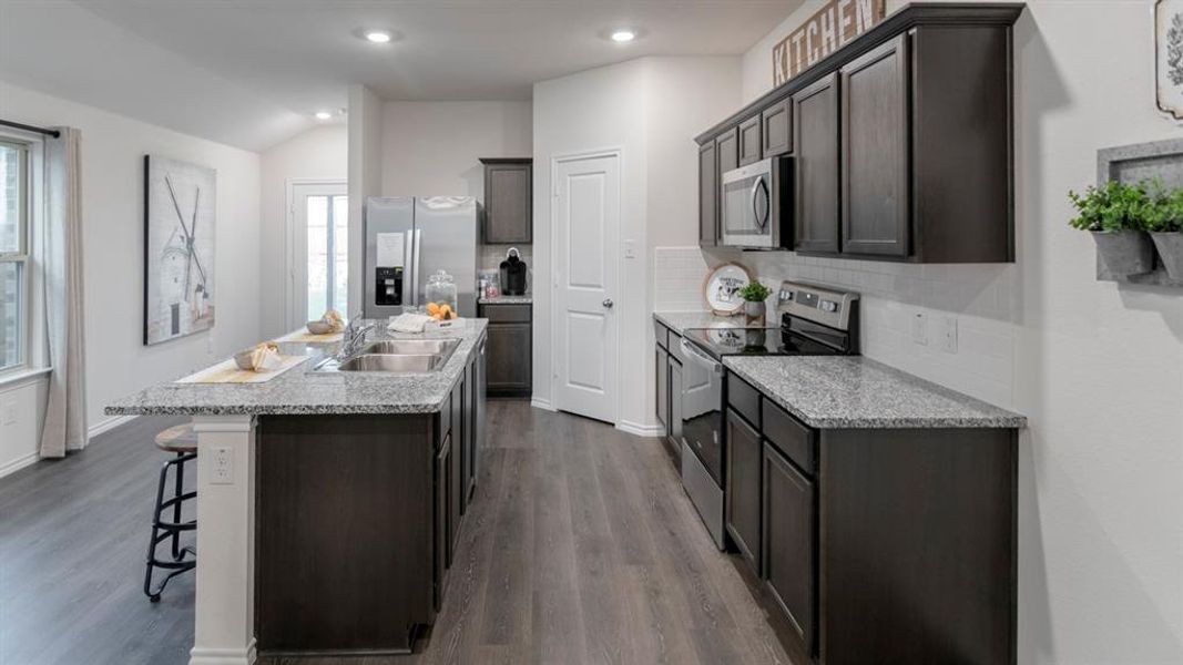 Open-concept kitchen featuring an expansive center island with dual sinks, light-toned stone-finish countertops, dark wood-finish cabinetry, stainless steel appliances, and wood-finish flooring Open-concept kitchen featuring an expansive center island with dual sinks, light-toned stone-finish countertops, dark wood-finish cabinetry, stainless steel appliances, and wood-finish flooring