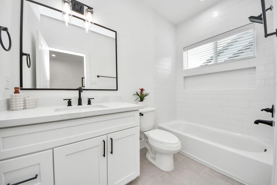 First floor bathroom features a modern design with a sleek white vanity, large mirror, black fixtures, and a bathtub with white subway tiles. Natural light streams in through a window with blinds, creating a bright and welcoming space.
