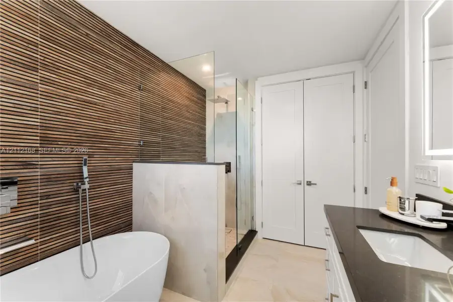 Bathroom featuring a textured wood-slat feature wall, freestanding soaking tub, walk-in glass-enclosed shower, dark countertop with an integrated basin, and double-door storage Bathroom featuring a textured wood-slat feature wall, freestanding soaking tub, walk-in glass-enclosed shower, dark countertop with an integrated basin, and double-door storage