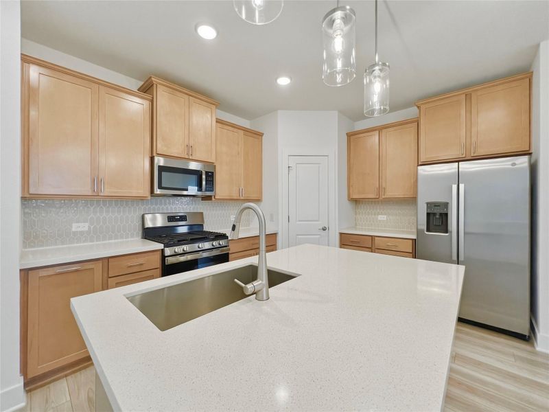 Kitchen with stainless steel appliances, backsplash, light wood finish cabinetry, a center island with sink, and hanging light fixtures