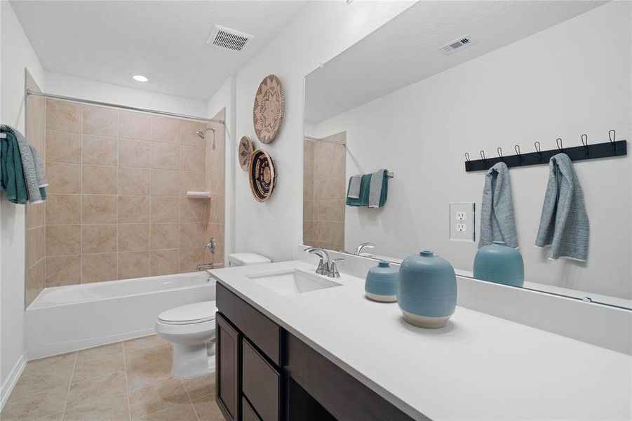 Secondary bath features tile flooring, bath/shower combo with tile surround, stained wood cabinets, beautiful light countertops, mirror, sleek and modern finishes.