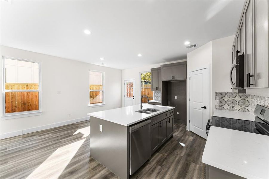 Kitchen featuring light stone counters, backsplash, stainless steel appliances, dark wood-style flooring, and a center island with sink Kitchen featuring light stone counters, backsplash, stainless steel appliances, dark wood-style flooring, and a center island with sink